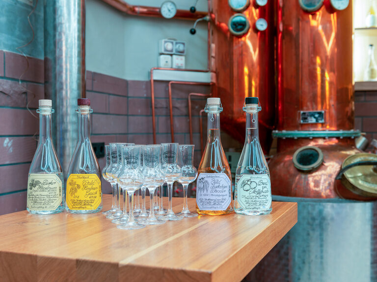 Auf einem Tisch vor großen Kesseln sind Gläser in einem schönen Dreieck aufgebaut und daneben stehen Flaschen.